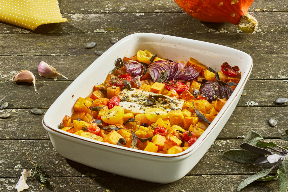 Gebackener K&uuml;rbis mit Feta, Kirschtomaten, roten Zwiebeln und Kr&auml;utern in wei&szlig;er Keramikauflaufform. Im Hintergrund ein K&uuml;rbis, Knoblauchzehen und Salbeibl&auml;tter auf rustikalem Holzuntergrund.