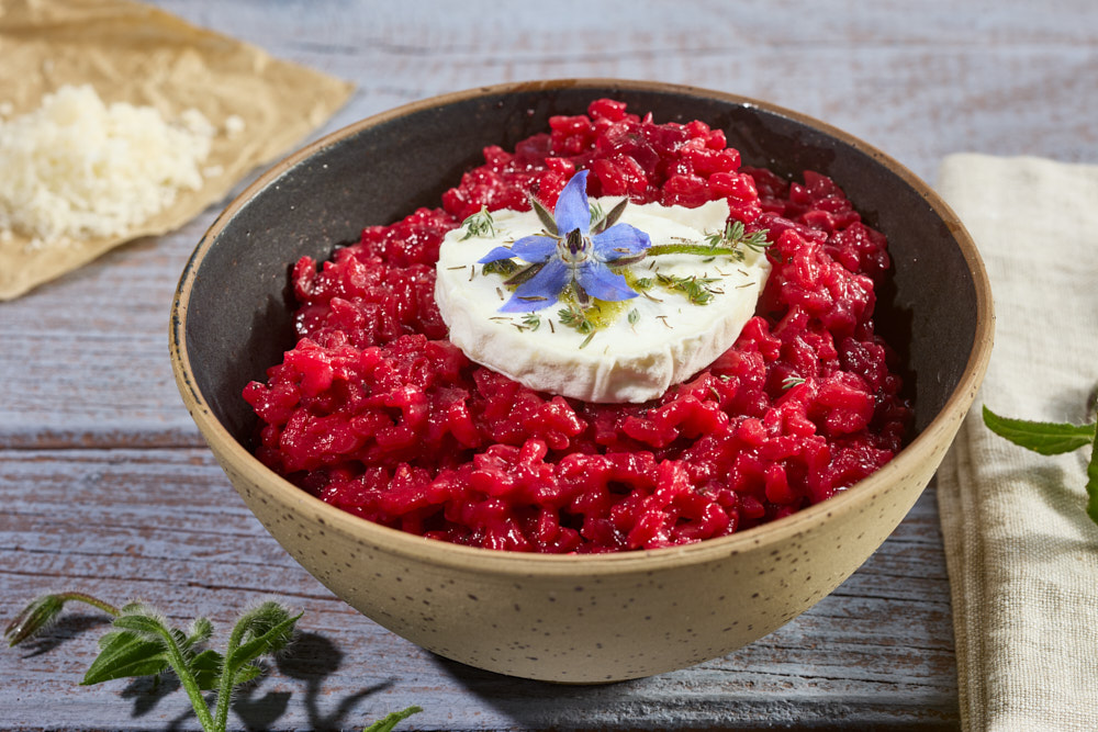 Rote Bete Risotto in dunkler Keramiksch&uuml;ssel, garniert mit Ziegen-Camembert, blauen Borretschbl&uuml;ten. Beigestellt: mit geriebenem Parmesan, Besteck auf Leinenserviette, Holzuntergrund.