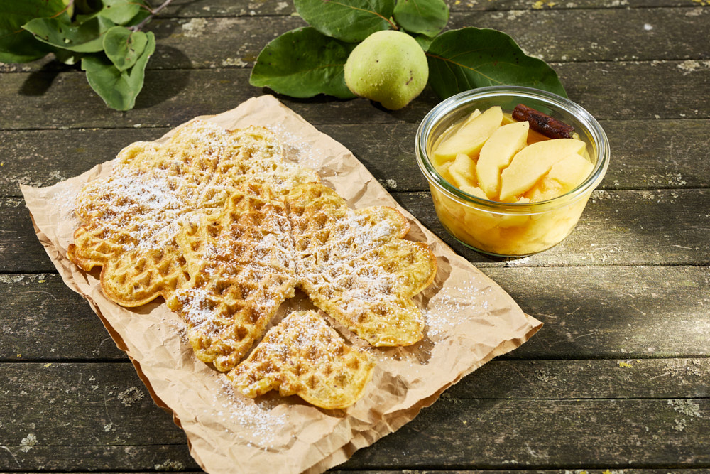 Herzf&ouml;rmige Nusswaffeln mit Puderzucker auf Backpapier, daneben ein Glasgef&auml;&szlig; mit Quittencompott. Im Hintergrund frische Quitten mit Bl&auml;ttern auf verwittertem Holztisch.